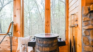 L’intérieur d’un chalet rustique en bois, avec une grande barrique en bois, une cheminée en pierre et une vue sur la forêt à travers de grandes fenêtres.