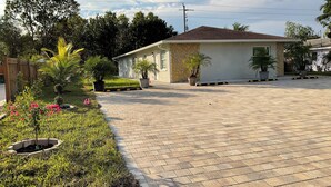 Une allée pavée mène à une maison dont la façade est en pierre et le toit recouvert de tuiles. On y trouve des plantes en pot et un petit jardin fleuri.