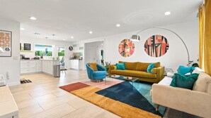 Una sala moderna con un sofá amarillo, un sillón azul y un sofá beige. Se observa una cocina al fondo con armarios blancos y un comedor.