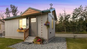 Una casa pequeña de un solo piso con un porche delantero, una escalera de madera y una cesta colgante con flores.