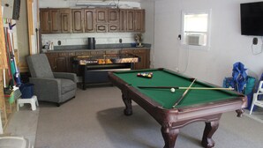 Een kamer met een pooltafel, een grijze fauteuil, houten kasten en een aan de muur bevestigde televisie.