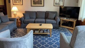 Een woonkamer met een grijze bank, een houten salontafel en een televisie op een standaard. Er hangen ingelijste foto’s aan de muur en staat een lamp op een bijzettafeltje.