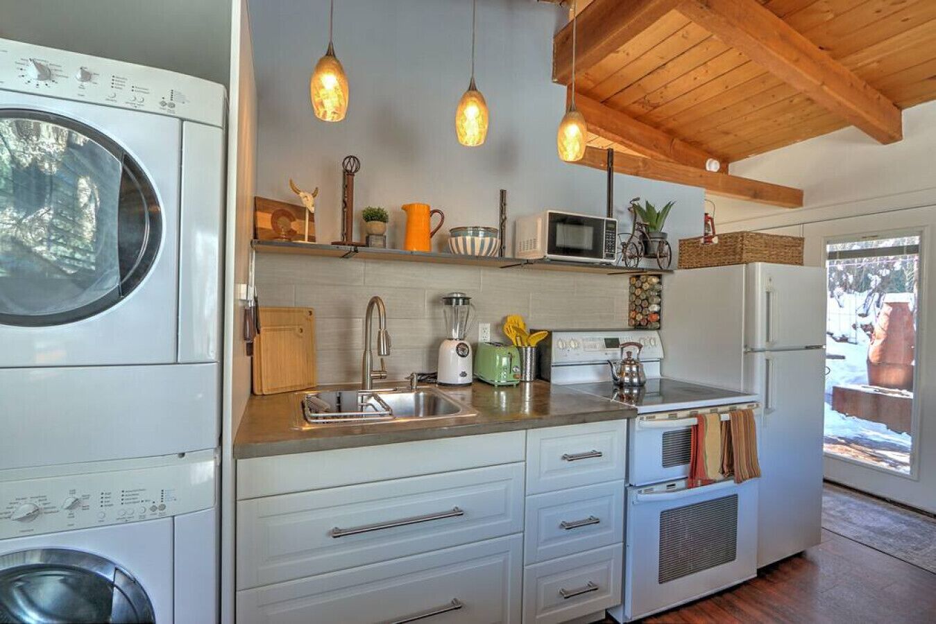 Una cucina compatta con lavatrice, microonde, fornello e lavandino. Sono presenti luci a sospensione, travi in legno al soffitto e una finestra con vista all'esterno.