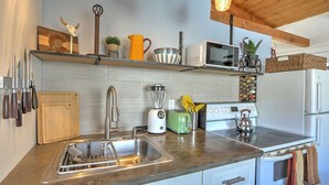 A modern kitchen with a stainless steel sink, a built-in toaster, a blender, and a microwave. There are wooden utensils hanging on the wall, and a shelf with decorative items.