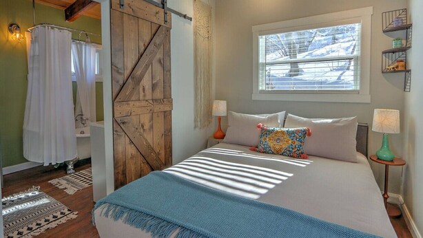 A bedroom with a bed, two bedside lamps, a window, a sliding barn door, and a shower area.