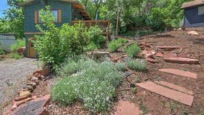 A rustic cabin with a wooden deck and a small porch, surrounded by a garden with various plants and a stone pathway.
