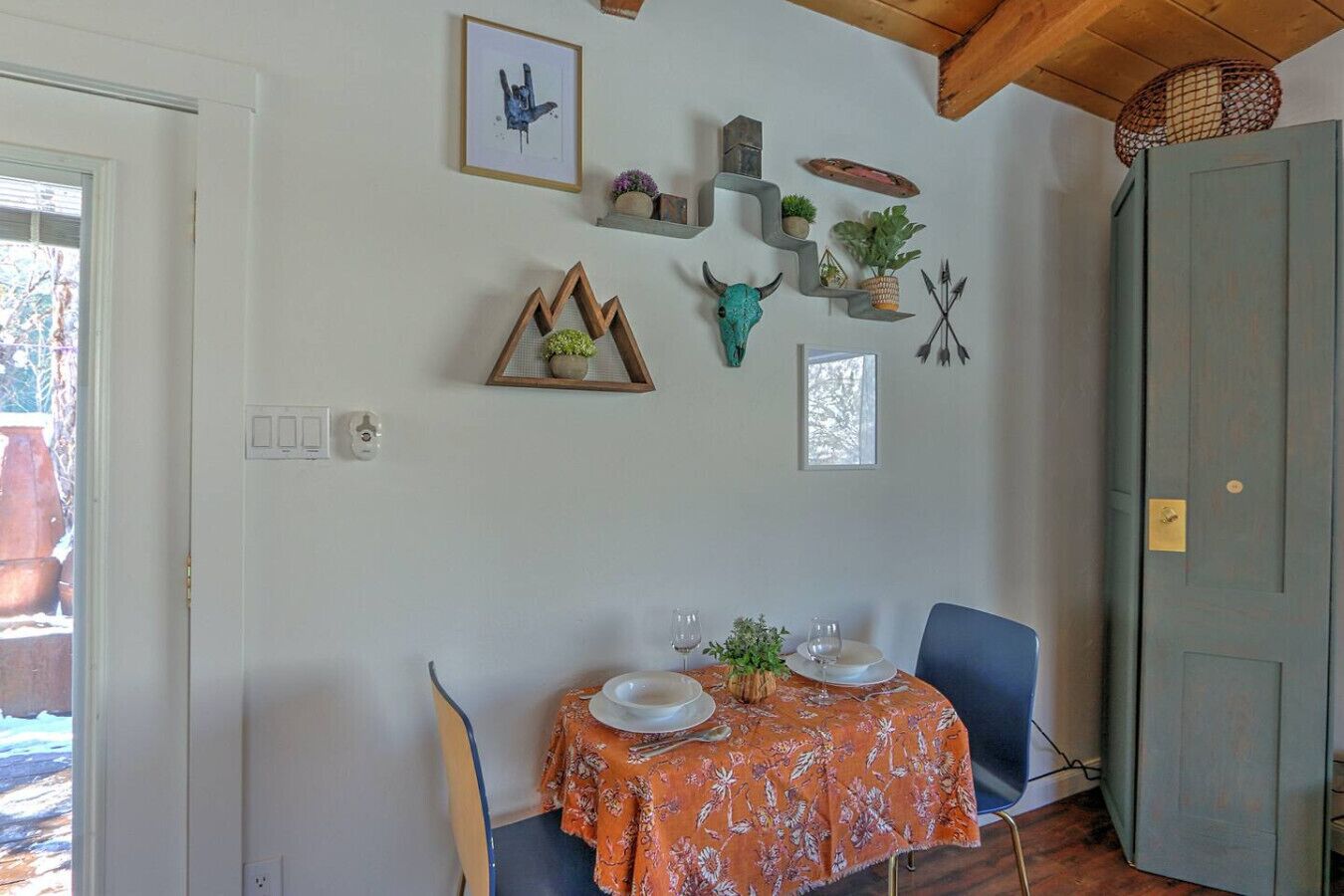 Een eetruimte met een tafel gedekt voor twee, versierd met een tafelkleed met bloemen. Er zijn wandplanken met diverse items, waaronder een ingelijst beeld, een klein plantje en een decoratieve schedel.