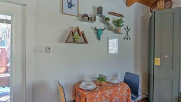 Een eetruimte met een tafel gedekt voor twee, versierd met een tafelkleed met bloemen. Er zijn wandplanken met diverse items, waaronder een ingelijst beeld, een klein plantje en een decoratieve schedel.