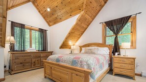 Un dormitorio con techo inclinado, muebles de madera, una cama con colcha estampada, dos mesitas de noche con lámparas y una ventana con cortinas.