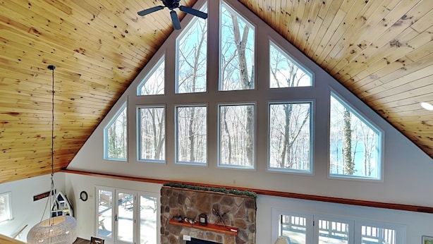 A spacious room with a high ceiling, large windows, a stone fireplace, and a ceiling fan. - Bearadise Found by Taylor Made Deep Creek (Swanton)