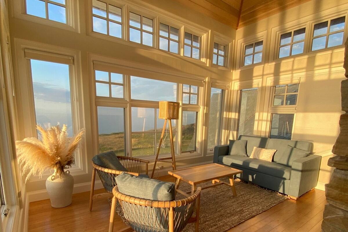 A spacious living room with large windows, a stone fireplace, and a view of the landscape.