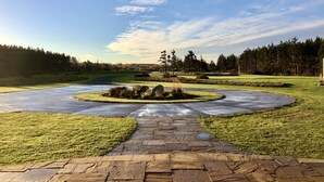 A paved pathway leading to a circular landscaped area with a small pond, surrounded by grass and trees. - 45 Steps - The Culinary Beachside Inn (Goose River)