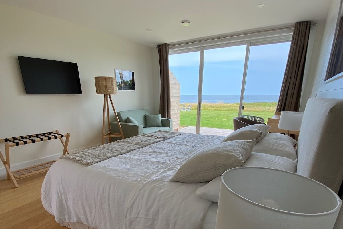 A bedroom with a large bed, a bedside table with a lamp, a TV on the wall, and a view of the sea through a sliding door.