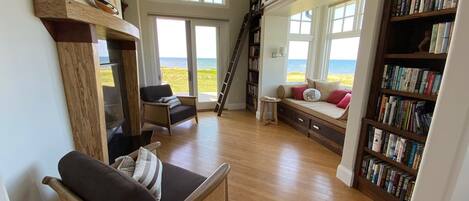 A spacious living room with a large bookshelf, a comfortable seating area with a view of the ocean, and a wooden floor.