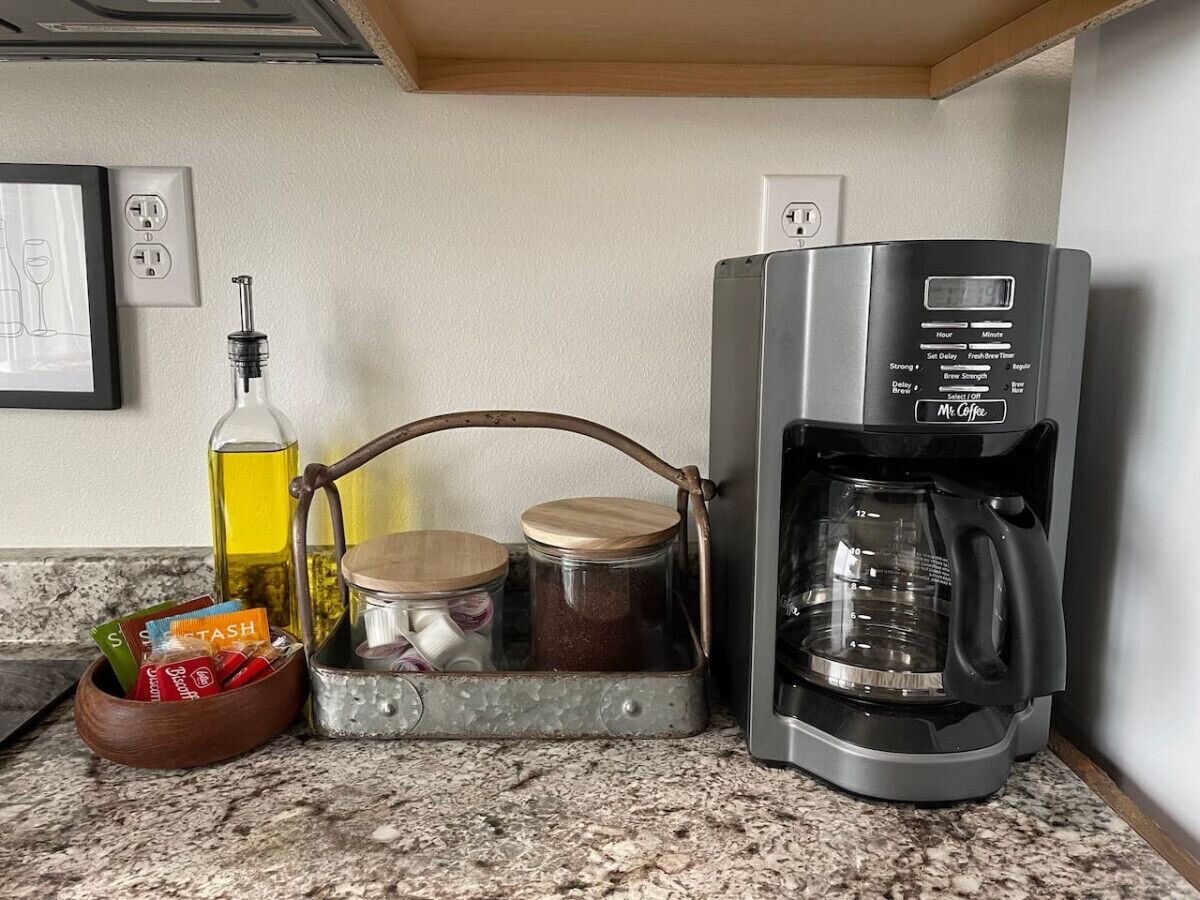 A kitchen counter with a coffee maker, a bottle of oil, a wooden bowl with snacks, and two jars.
