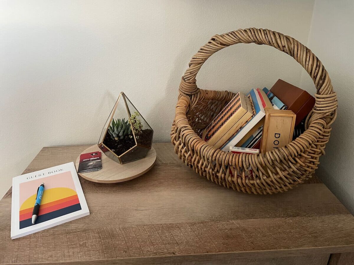 Une table en bois sur laquelle se trouvent un registre pour les clients, un terrarium en verre et un panier en osier contenant des livres et une petite plante.