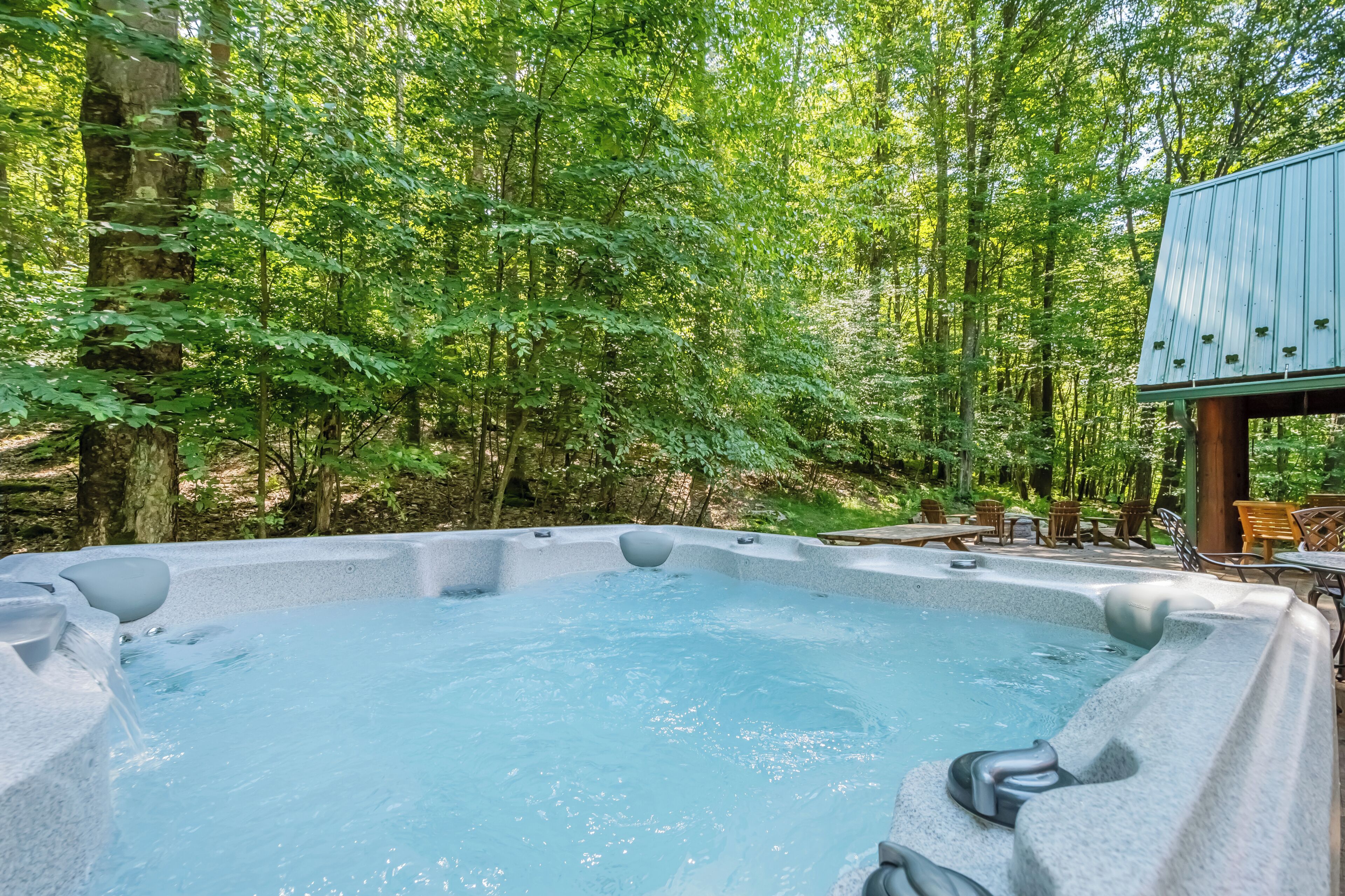 A hot tub surrounded by a forested area with a wooden deck and chairs.