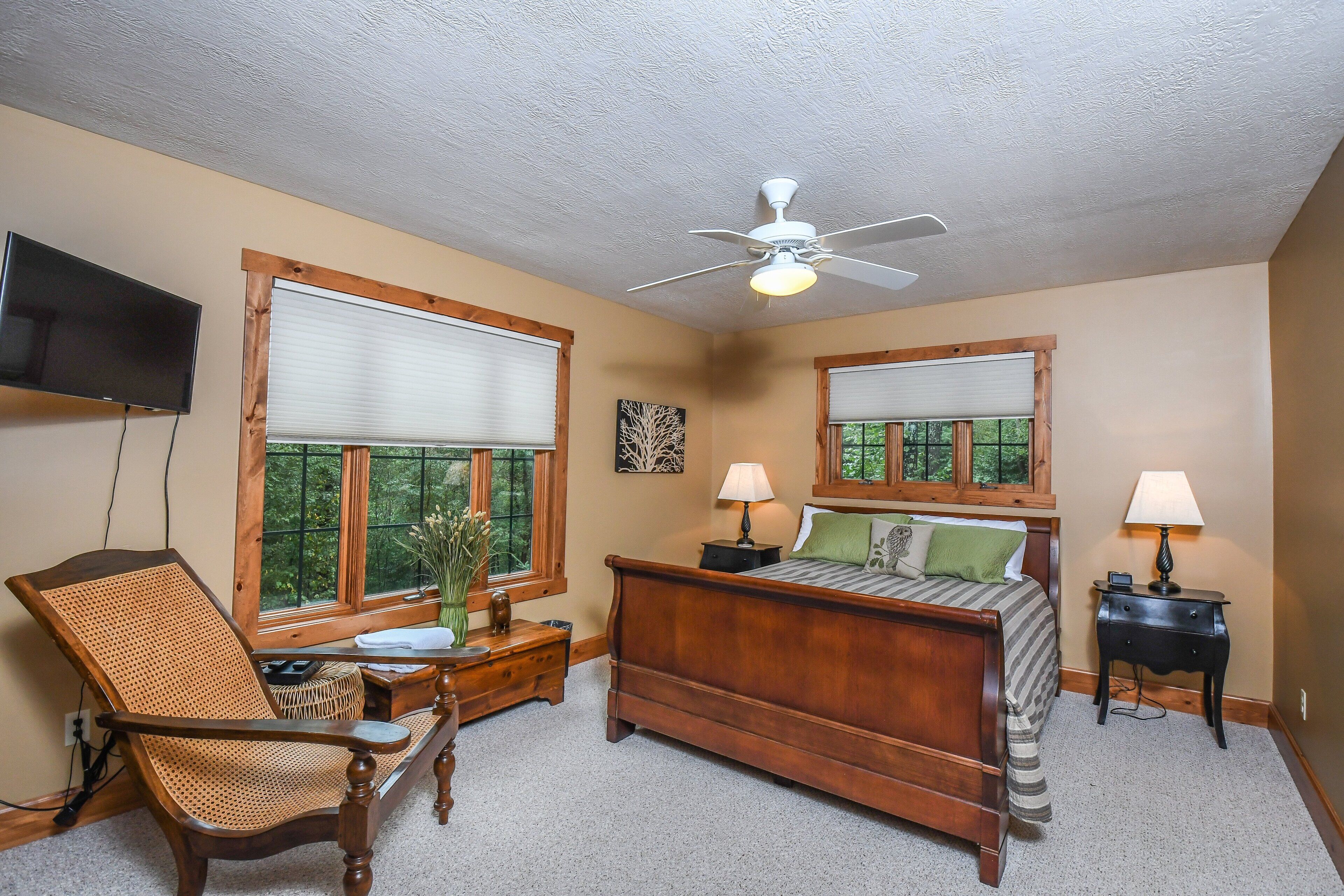 A bedroom with a wooden bed, two bedside tables with lamps, a chair, a television, and large windows with blinds.