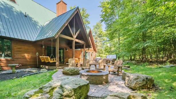 A rustic cabin with a metal roof, a stone fire pit, and wooden furniture in a forested area.