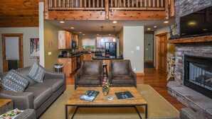 A spacious living room with a high ceiling, a fireplace, a wooden staircase, and a kitchen in the background.
