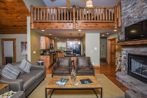 A spacious living room with a high ceiling, a fireplace, a wooden staircase, and a kitchen in the background.