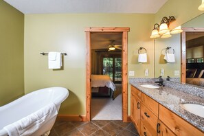 A bathroom with a bathtub, a sink, and a mirror. There is a door leading to a bedroom.