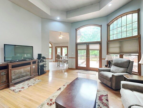 A spacious living room with a flat-screen TV, a wooden entertainment unit, a large window with a view of the outdoors, and a comfortable seating area with a sofa and armchairs. - Beautiful Adventure by Taylor Made Deep Creek (Swanton)