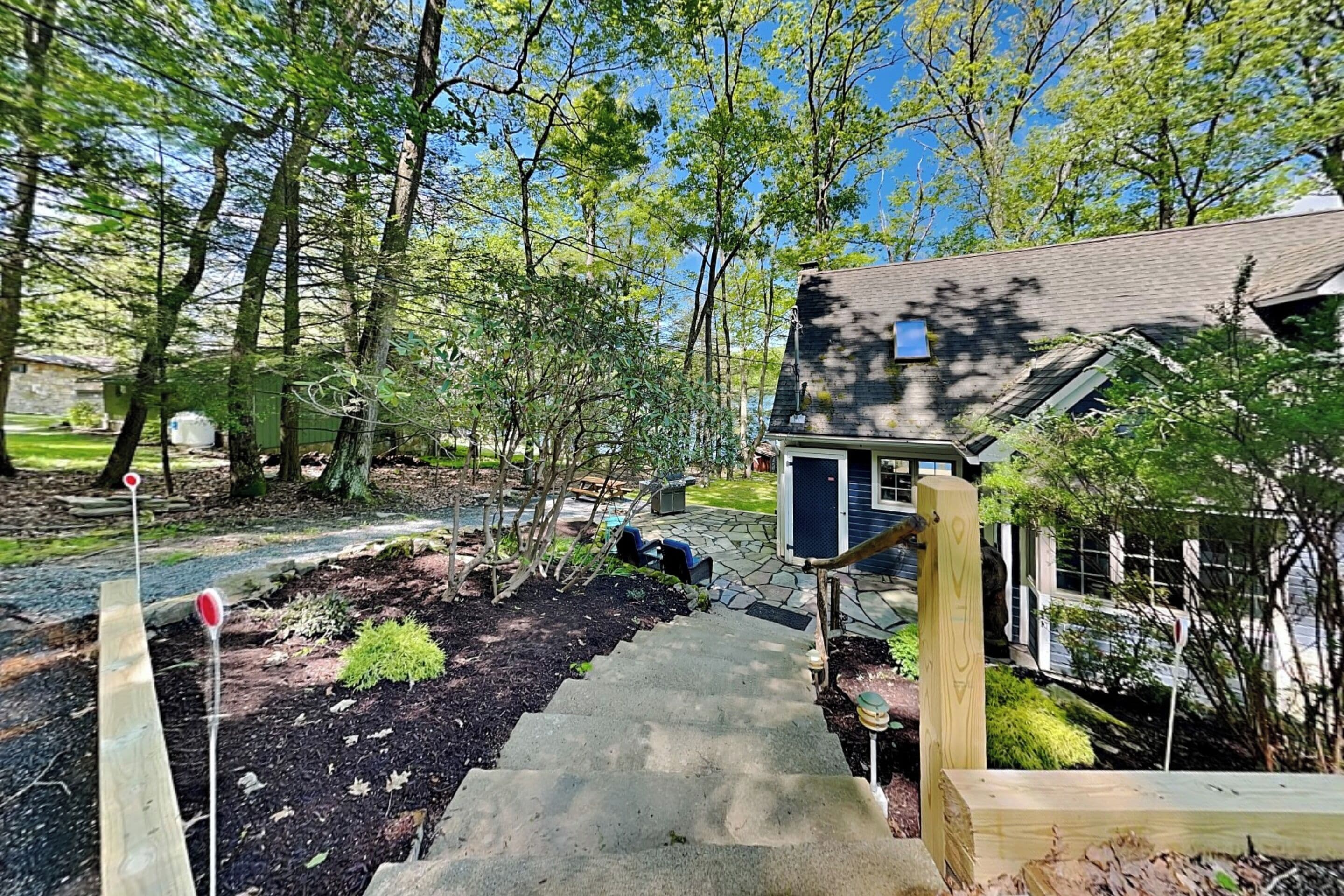 Une terrasse en bois mène à une maison, un chemin de pierres la longe, et l’ensemble est entouré d’arbres et d’un jardin.