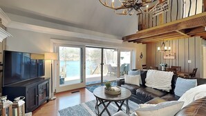 A living room with a TV, leather sofa, and a view of the lake. - Bearfoot Lodge by Taylor Made Deep Creek (Oakland)
