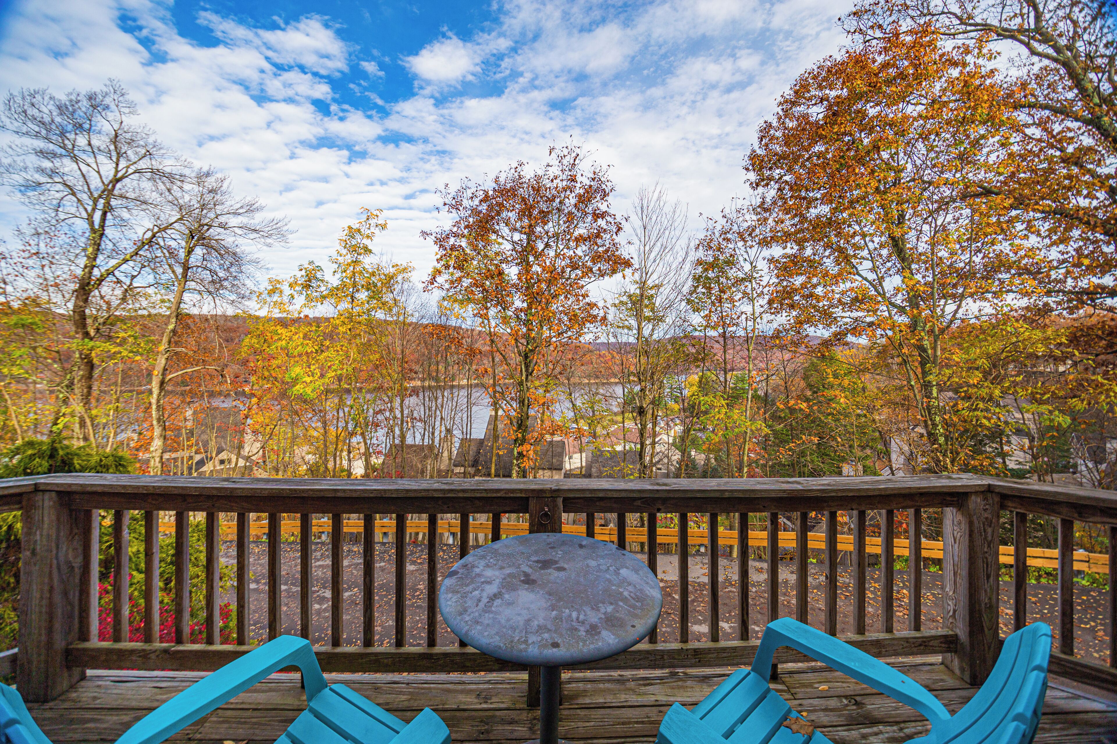 A wooden deck with a round table and blue chairs, overlooking a scenic autumn landscape with trees and a distant building.