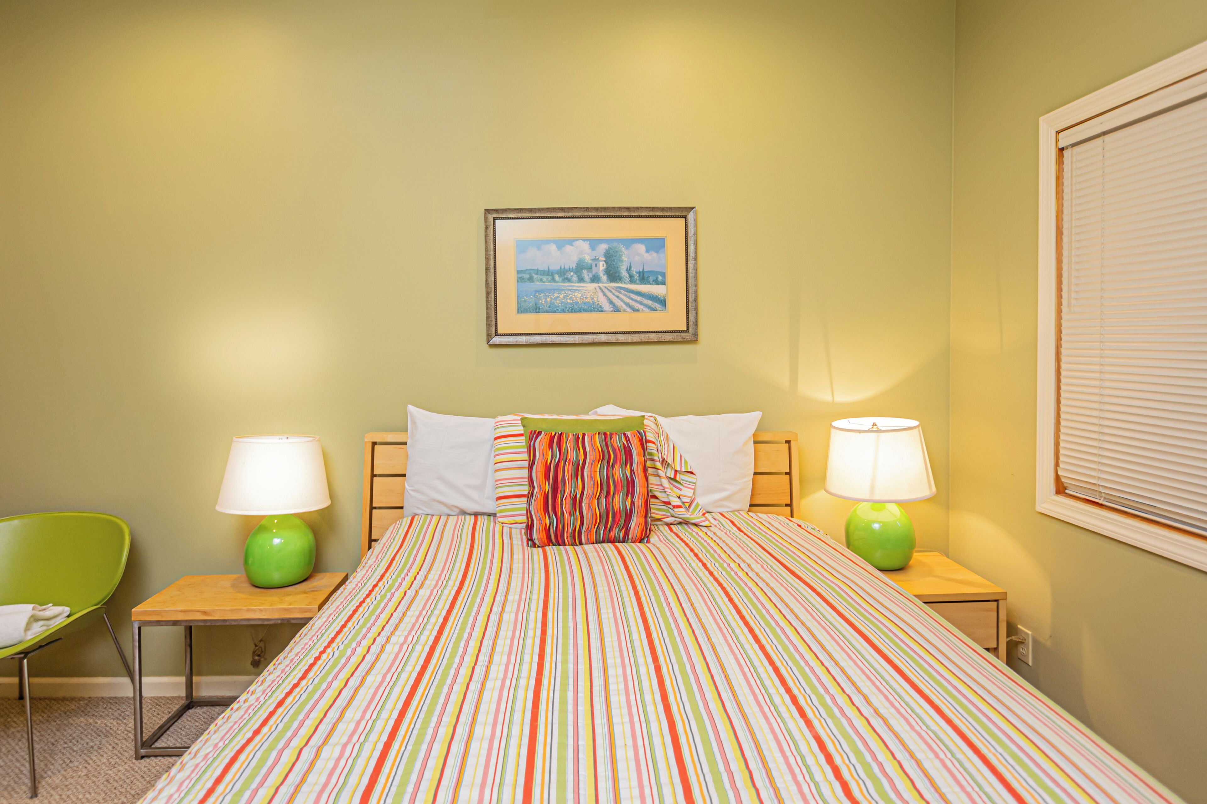 Habitación de hotel con cama, dos mesitas de noche, dos lámparas, una silla verde, un cuadro en la pared y una ventana con persianas.
