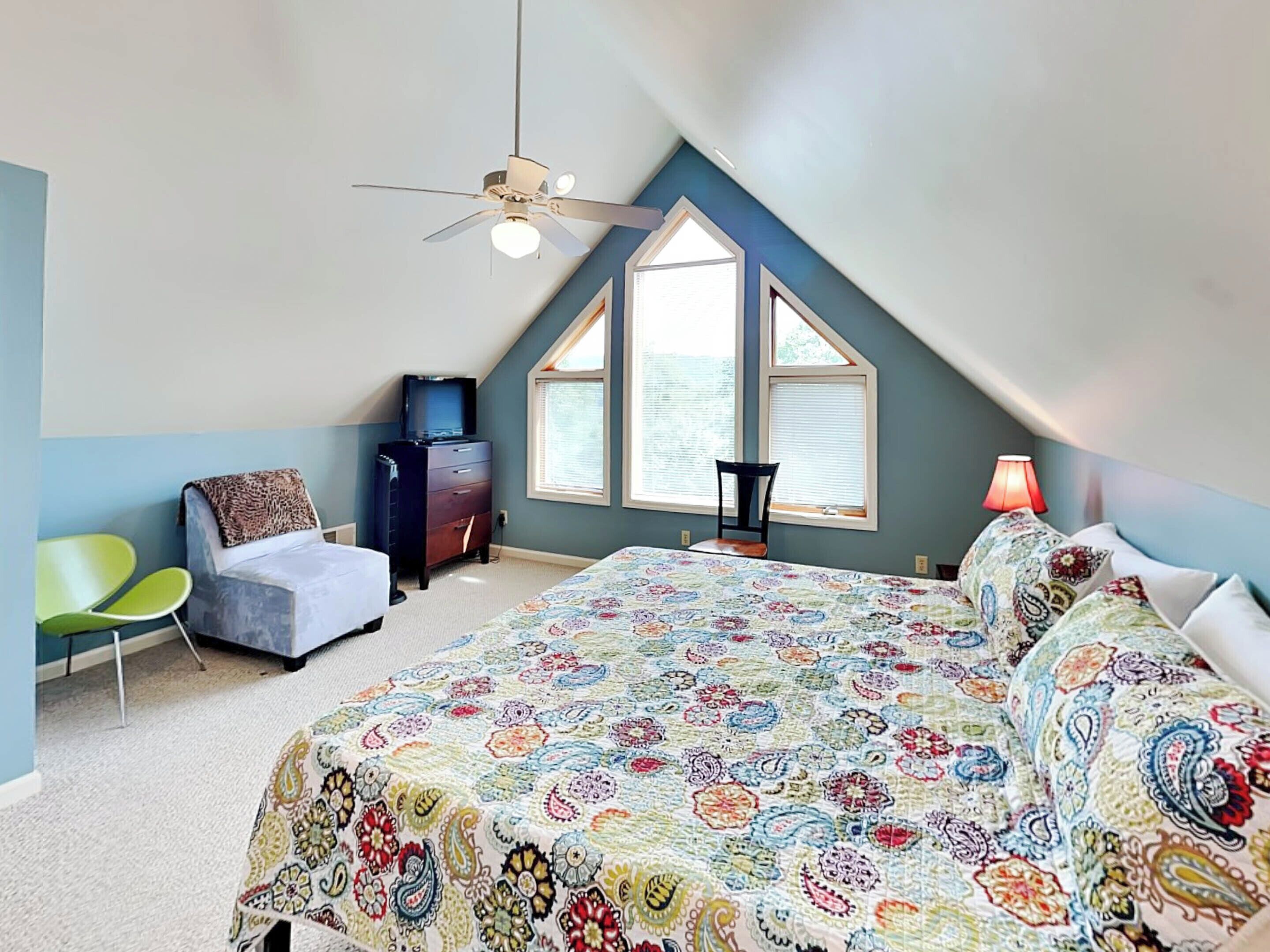 A bedroom with a sloped ceiling, a bed with a colorful floral bedspread, a chair, a dresser, and a ceiling fan.
