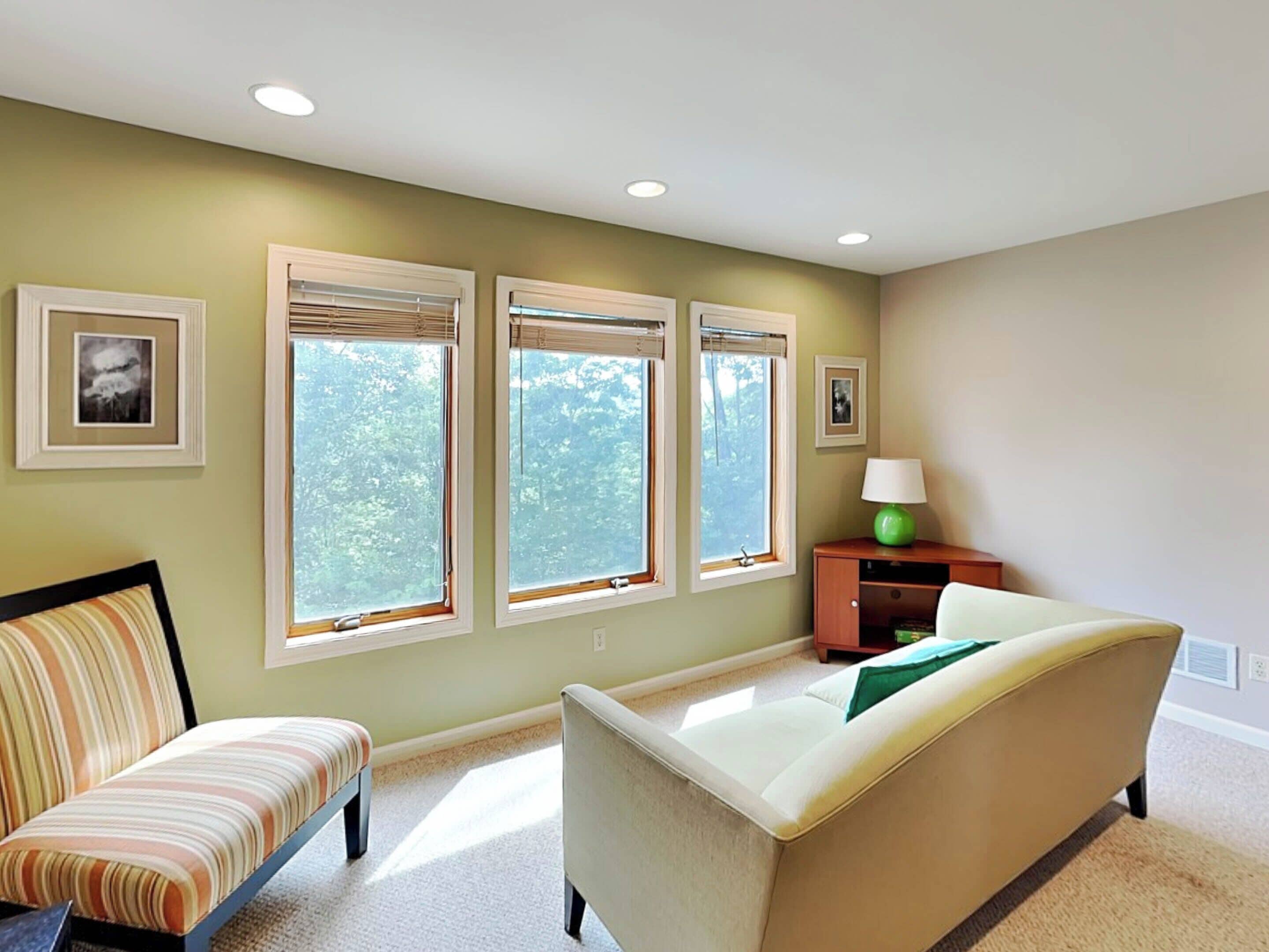 Una habitación con tres ventanas amplias, un sofá, una silla y una lámpara sobre una mesita auxiliar.