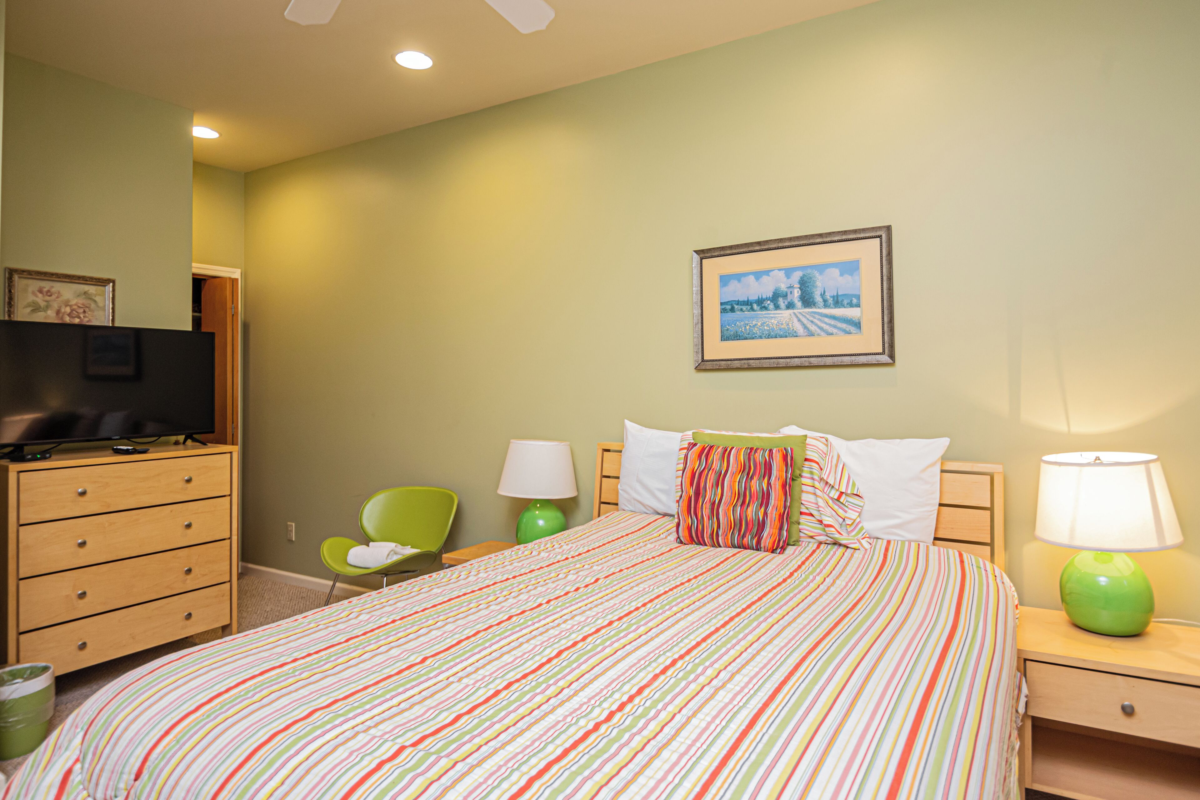 A hotel room with a bed, a television, a green chair, a nightstand, and a lamp.