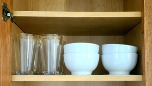 Un mueble de madera con estantes que contienen vasos transparentes, cuencos blancos y platos blancos apilados.