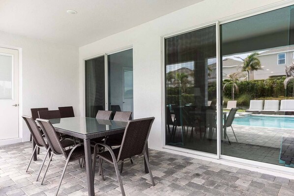 A modern outdoor dining area with a glass table and chairs, a pool, and a view of a residential neighborhood.