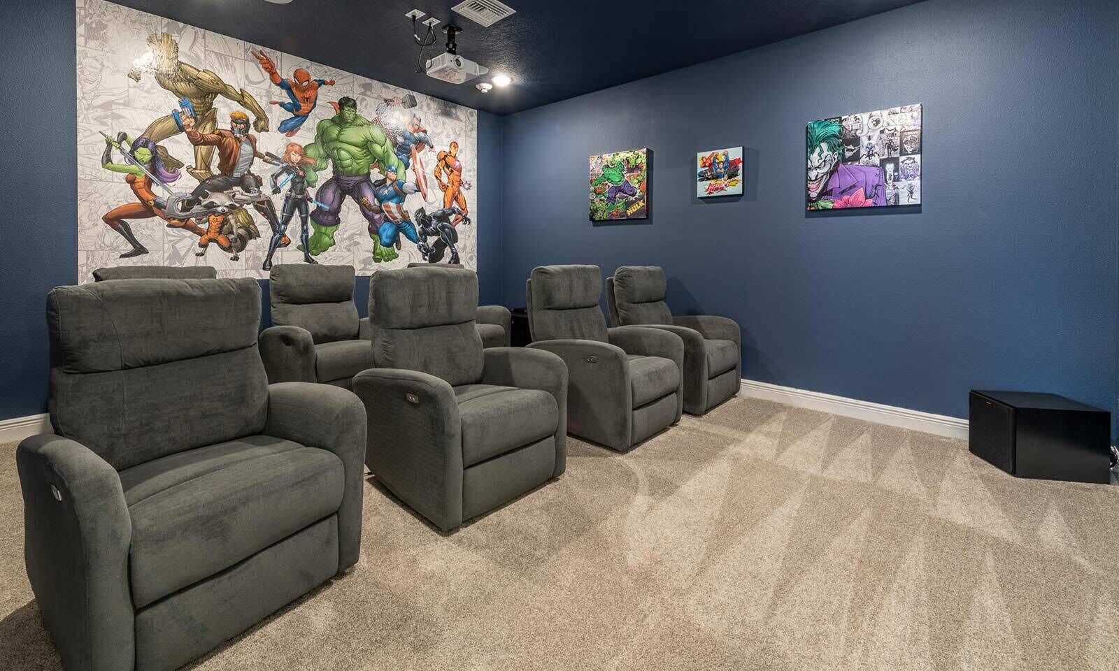 A home theater room with six gray reclining chairs, a large wall mural of comic book characters, and three framed comic book covers on the wall.
