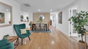 A spacious living room with a wooden floor, a dining area with a round table and chairs, and a large mirror on the wall.