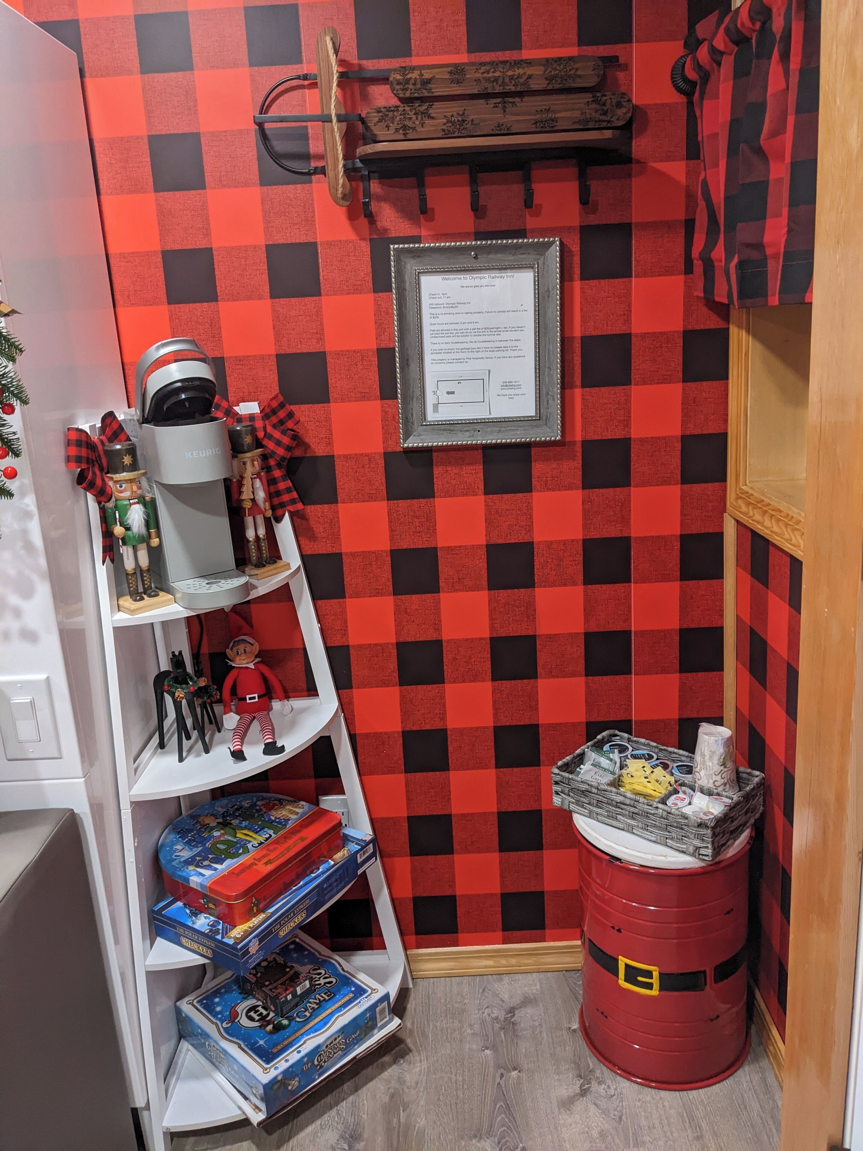 A small, cozy room with a red and black checkered wall, a white shelf with toys, a red barrel, and a framed welcome sign.