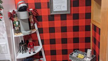 A small, cozy room with a red and black checkered wall, a white shelf with toys, a red barrel, and a framed welcome sign.