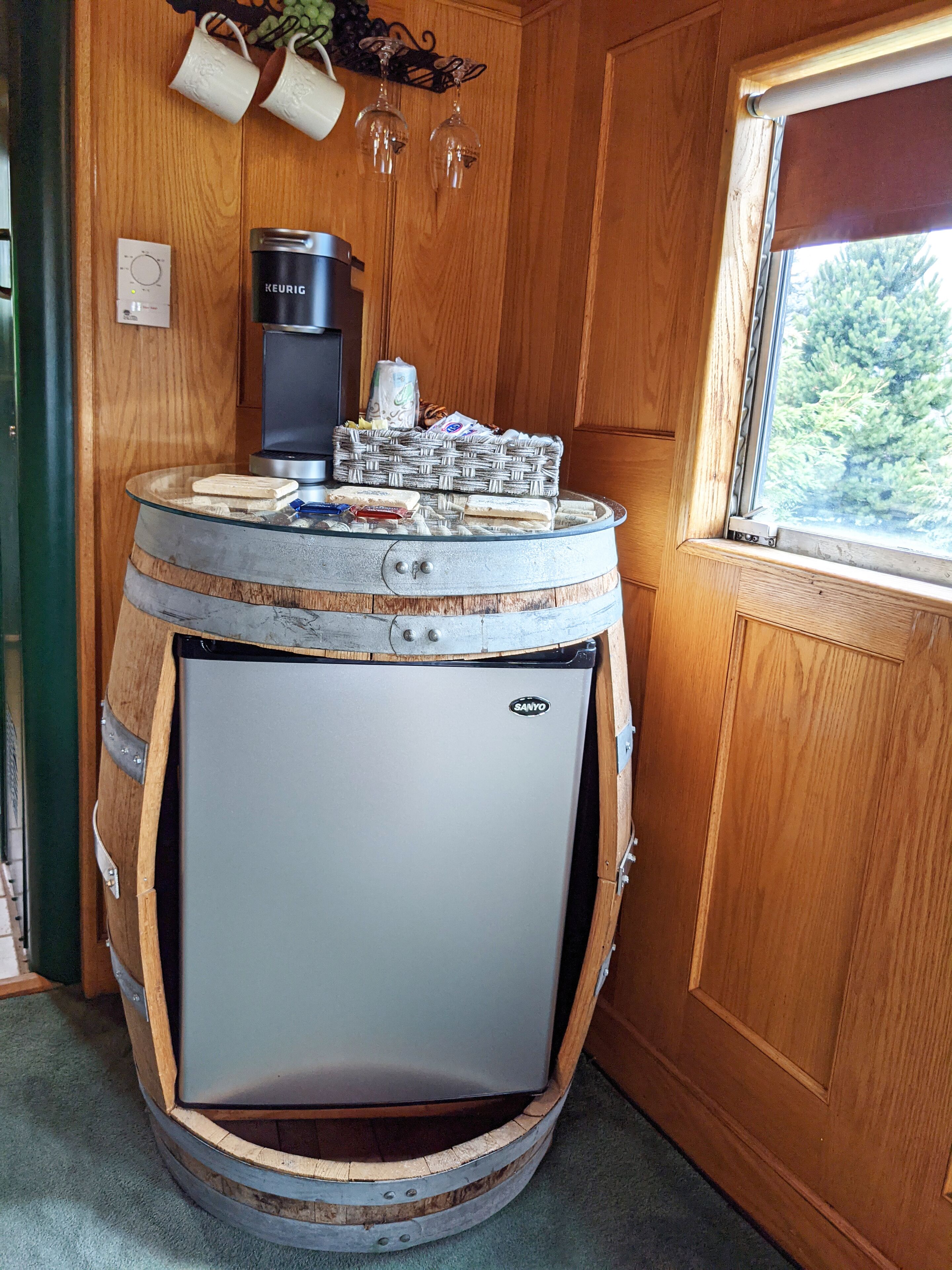 Un refrigerador compacto dentro de un barril de madera, una cafetera y una cesta de mimbre con aperitivos encima.
