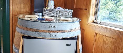 Un refrigerador compacto dentro de un barril de madera, una cafetera y una cesta de mimbre con aperitivos encima.