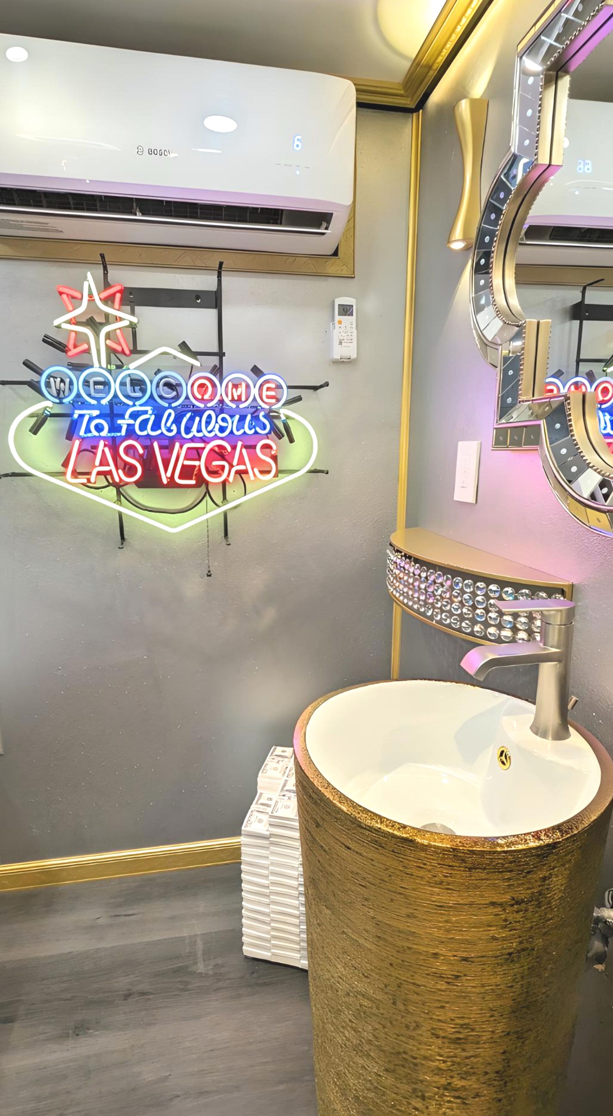 Et badeværelse på et hotel med et neonskilt, der siger "Welcome to Fabulous Las Vegas", et stort spejl med lys og en rund vask med dekorativ fod.