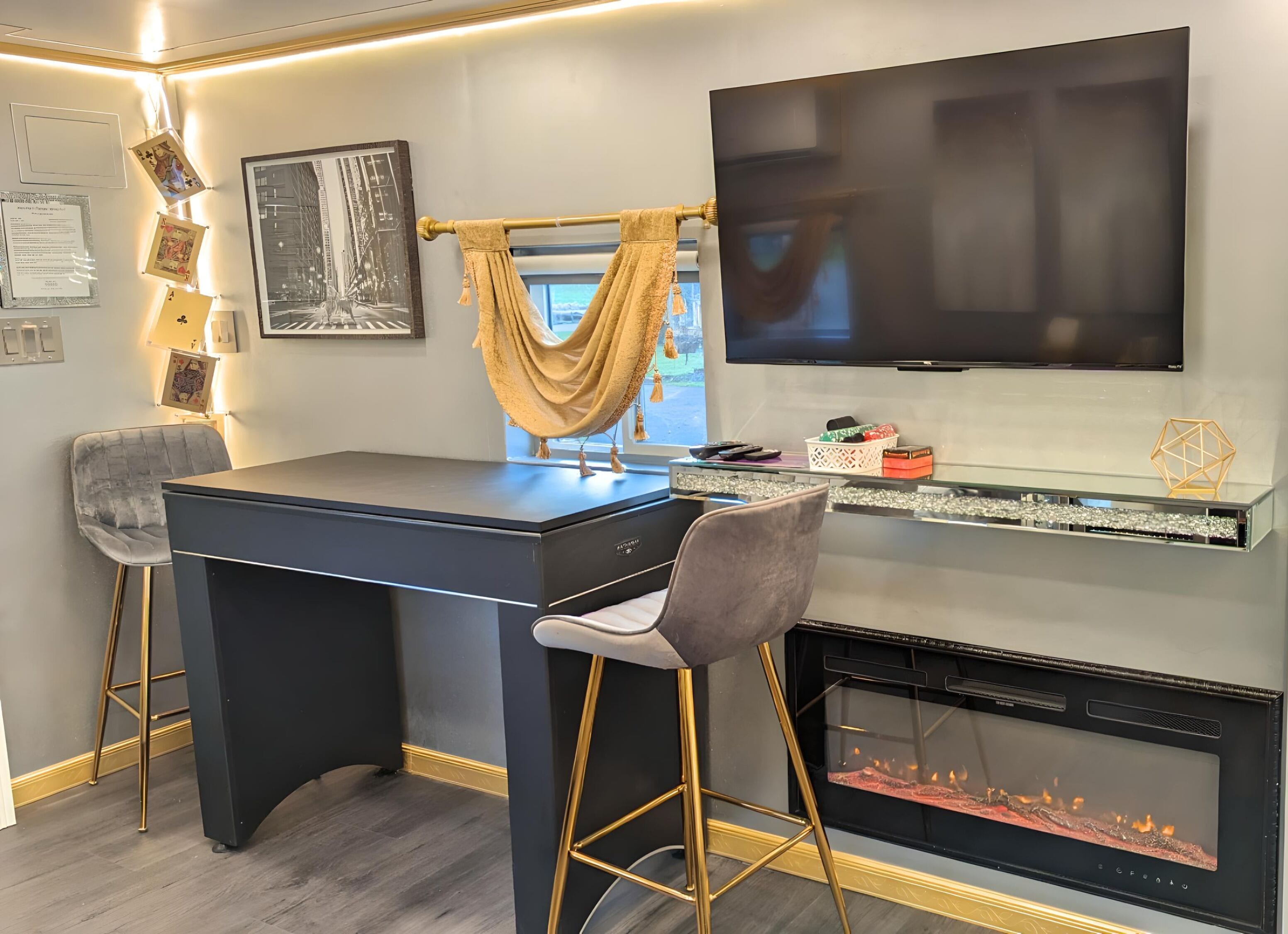 A modern living room with a dark-colored desk, two bar stools, a flat-screen TV mounted on the wall, a fireplace, and a window with a view of the outdoors.