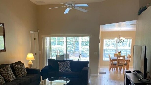 A living room with a sofa, coffee table, and a TV stand. There is a ceiling fan and a window with a view of greenery outside. - Cozy 3Be/2Ba Home, Heated Pool/Spa, near Orlando-Disney, Universal, Golf Course. (Davenport)