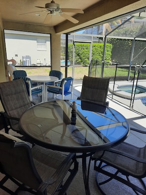 A sunlit patio with a glass table and chairs, a ceiling fan, and a view of a backyard.