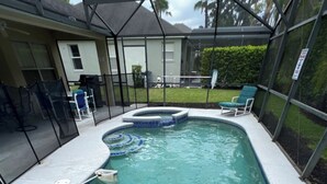 A swimming pool with a curved design, surrounded by a covered patio area with chairs and a small hot tub. - Cozy 3Be/2Ba Home, Heated Pool/Spa, near Orlando-Disney, Universal, Golf Course. (Davenport)