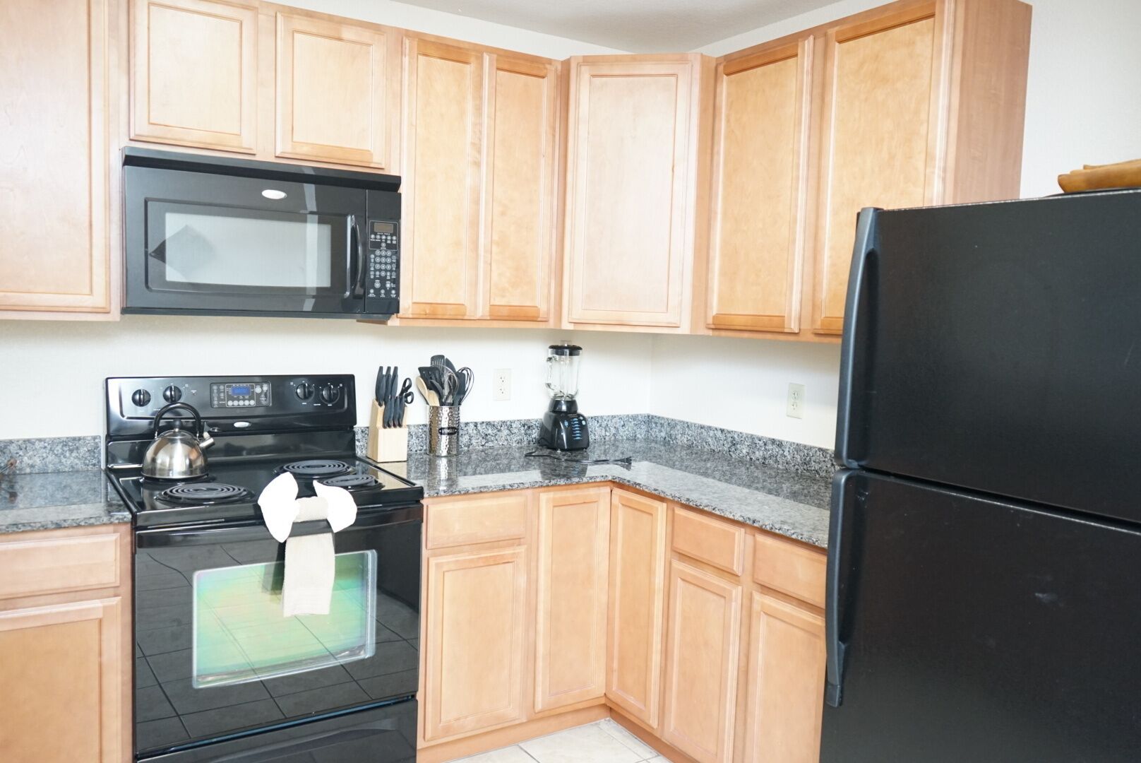Una cocina con armarios de madera, estufa negra, microondas, refrigerador y hervidor.