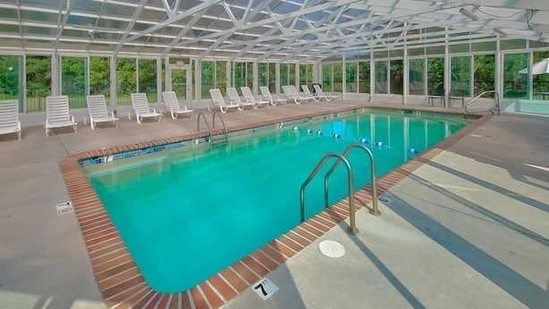 An indoor swimming pool with clear blue water, surrounded by white lounge chairs and a glass ceiling structure.