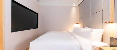 A modern hotel room with a large bed, a flat-screen TV mounted on the wall, and a bedside table with a lamp.